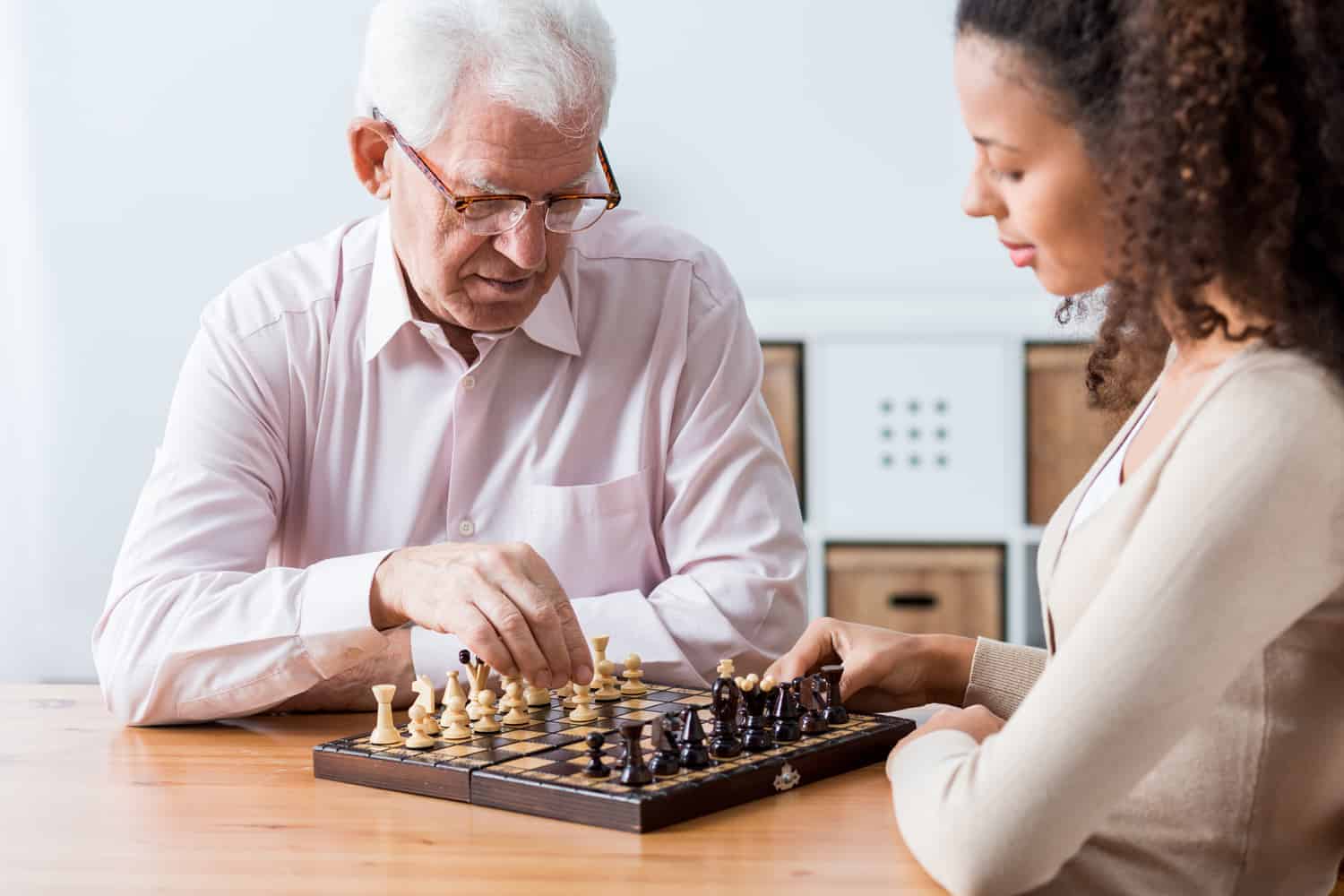 An elderly man and a young woman thoughtfully play chess together at a wooden table, sharing moments before moving a loved one to a care home.