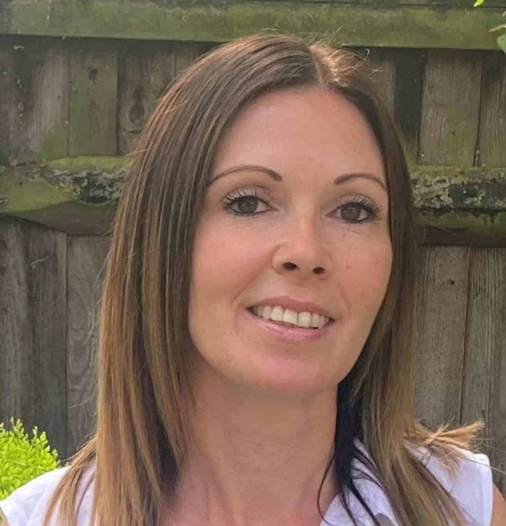 A smiling woman with straight brown hair, part of our team, standing in front of a wooden fence and greenery.