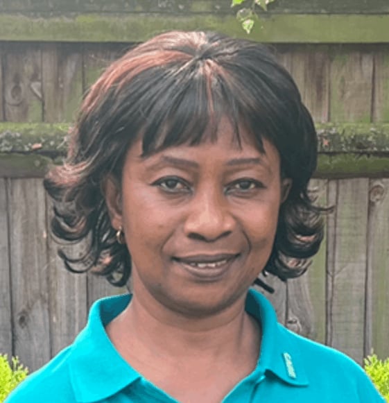 A portrait of a smiling woman with short curly hair, wearing a turquoise shirt, part of the team, standing in front of a wooden fence with greenery peeking out.