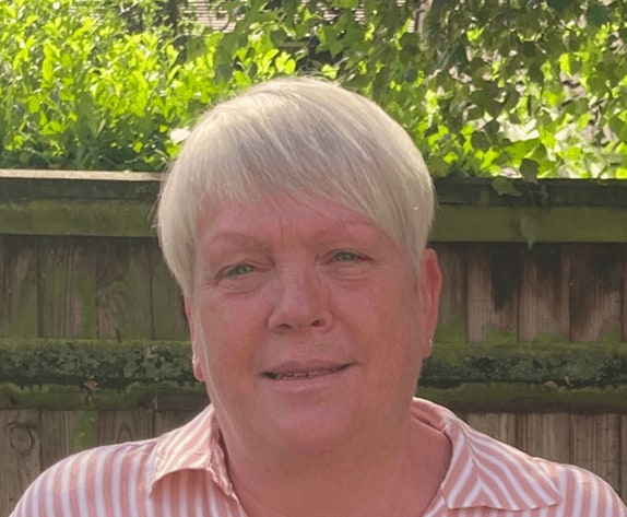 A person with short blonde hair, wearing a pink and white striped shirt, smiles slightly in front of a green foliage backdrop and a wooden fence as part of our meet the team photo session.