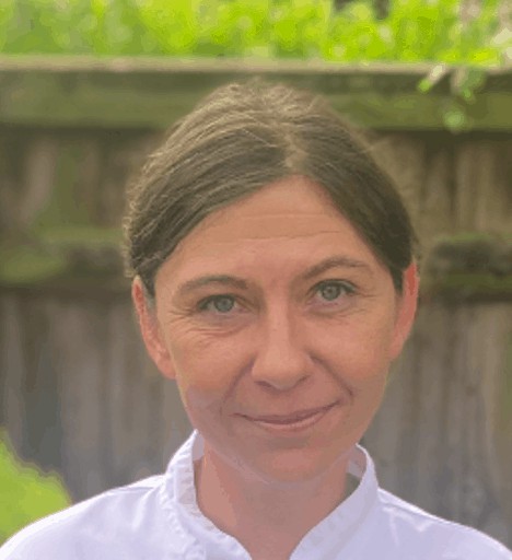A woman with a subtle smile standing in front of a wooden fence with green foliage in the background, ready to meet the team.