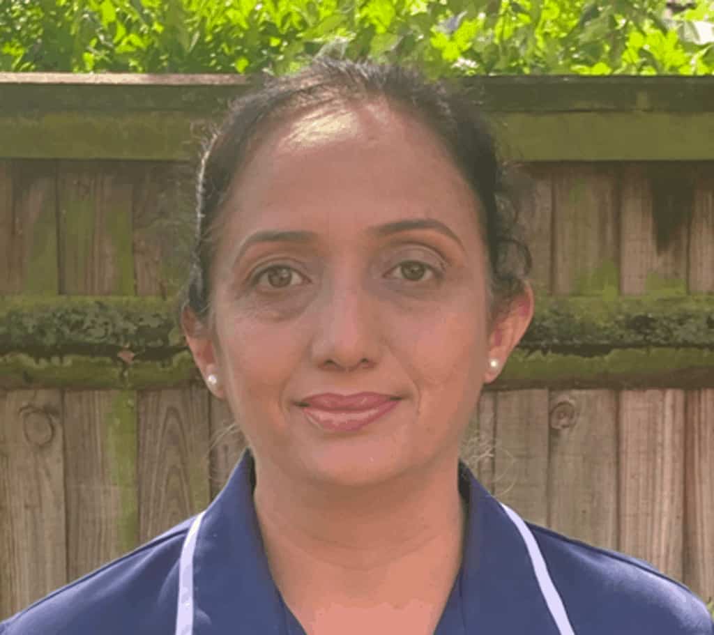 A woman with a gentle smile standing outdoors with a wooden fence and green foliage in the background, ready to meet the team.
