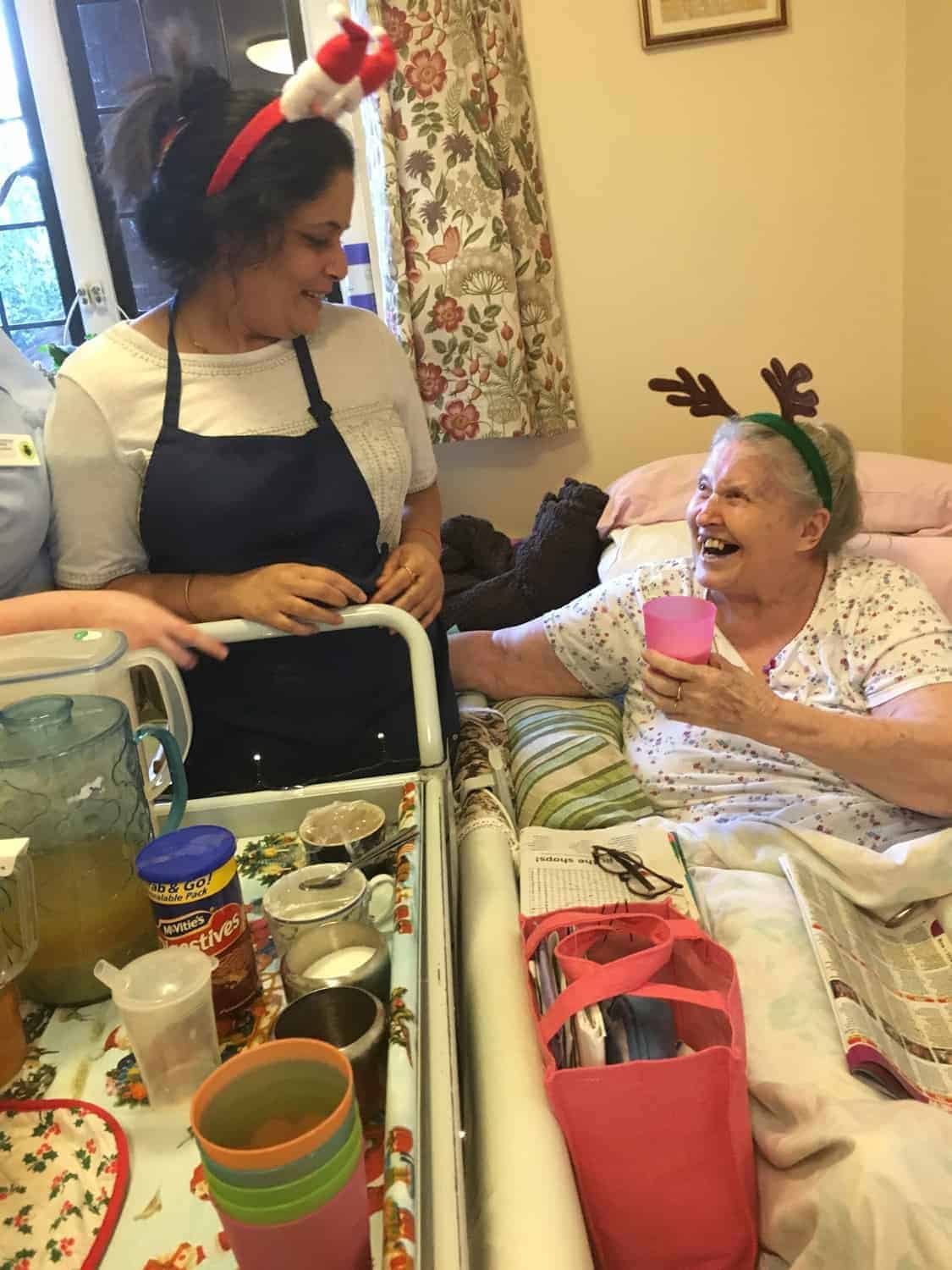 A heartwarming moment in a gallery with two individuals celebrating the holiday spirit, as a younger woman wearing a festive hat shares a joyful conversation with an elderly woman sporting reindeer antlers, who is