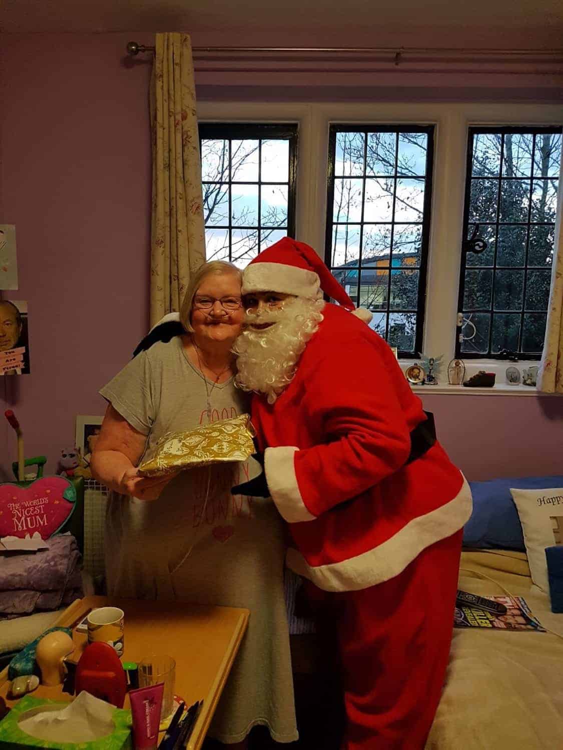 A festive moment as a woman shares a smile with someone dressed as Santa Claus while holding a gift, celebrating the holiday spirit in the cozy room of an art gallery.
