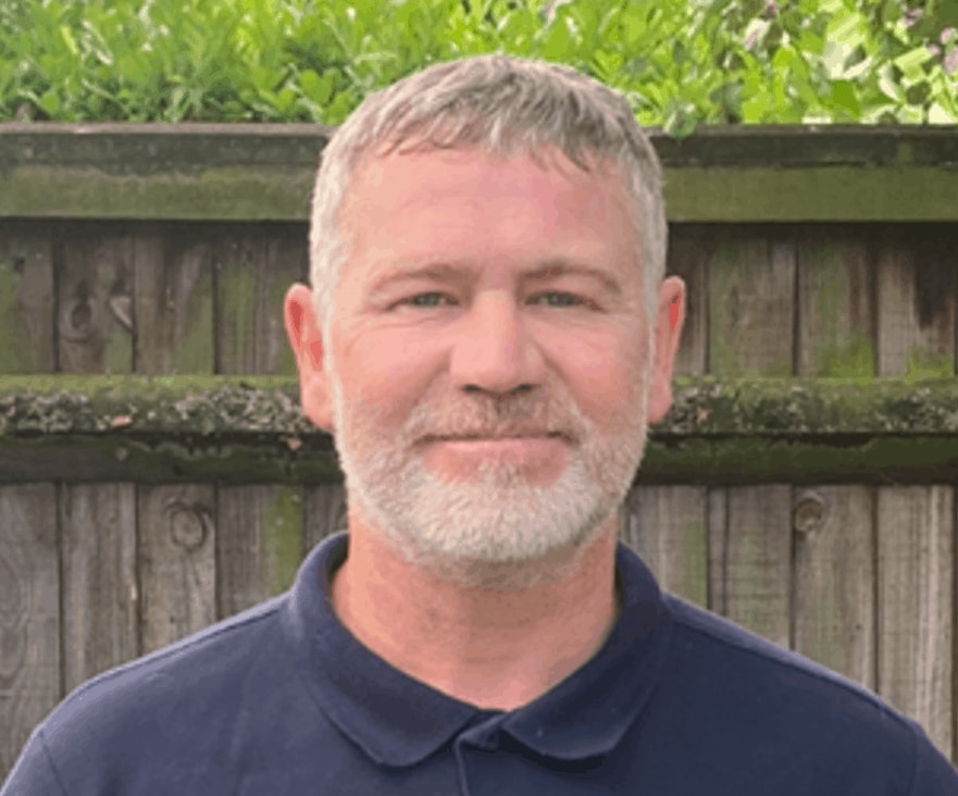 A smiling man with a greying beard, short hair, and a dark polo shirt poses in front of a wooden fence with green foliage in the background for his "meet the team" photo.