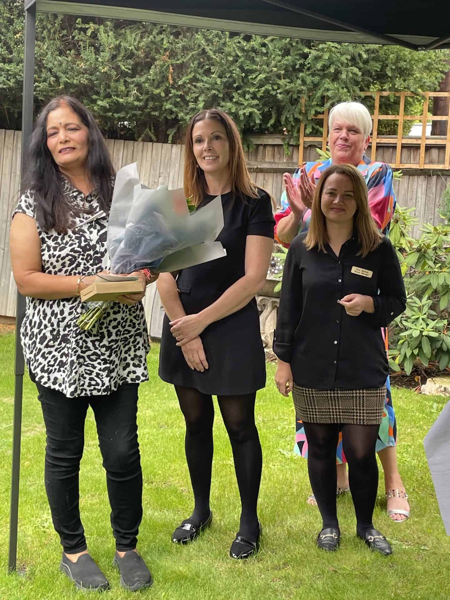 A group of four women in a garden, with one holding a bouquet of flowers, smiling at the camera during a daytime gathering or event.