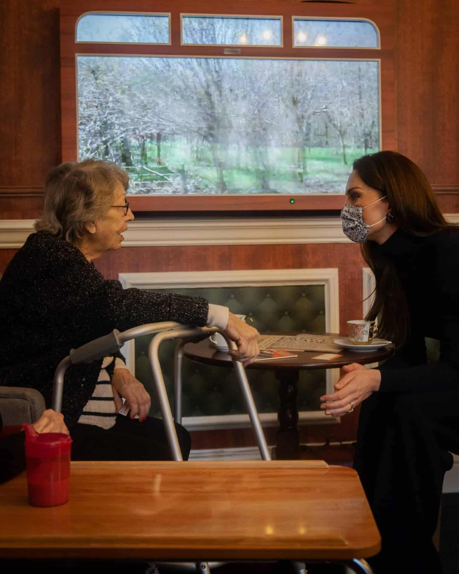 A heartfelt conversation indoors at the Oxford House Nursing Home in Slough: an elderly woman seated with a walker interacts with a younger woman wearing a face mask; a tranquil garden visible through the window in the