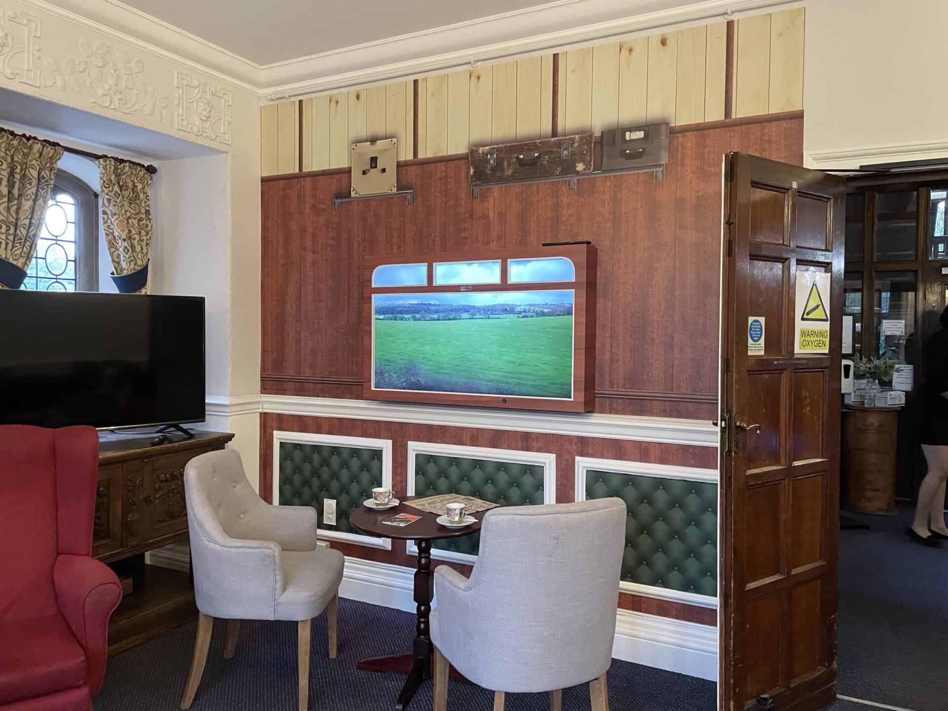 A cozy seating area in a traditional room within Oxford House Nursing Home, featuring comfortable armchairs, a small table set for tea, and an interactive feature displaying a scenic landscape.