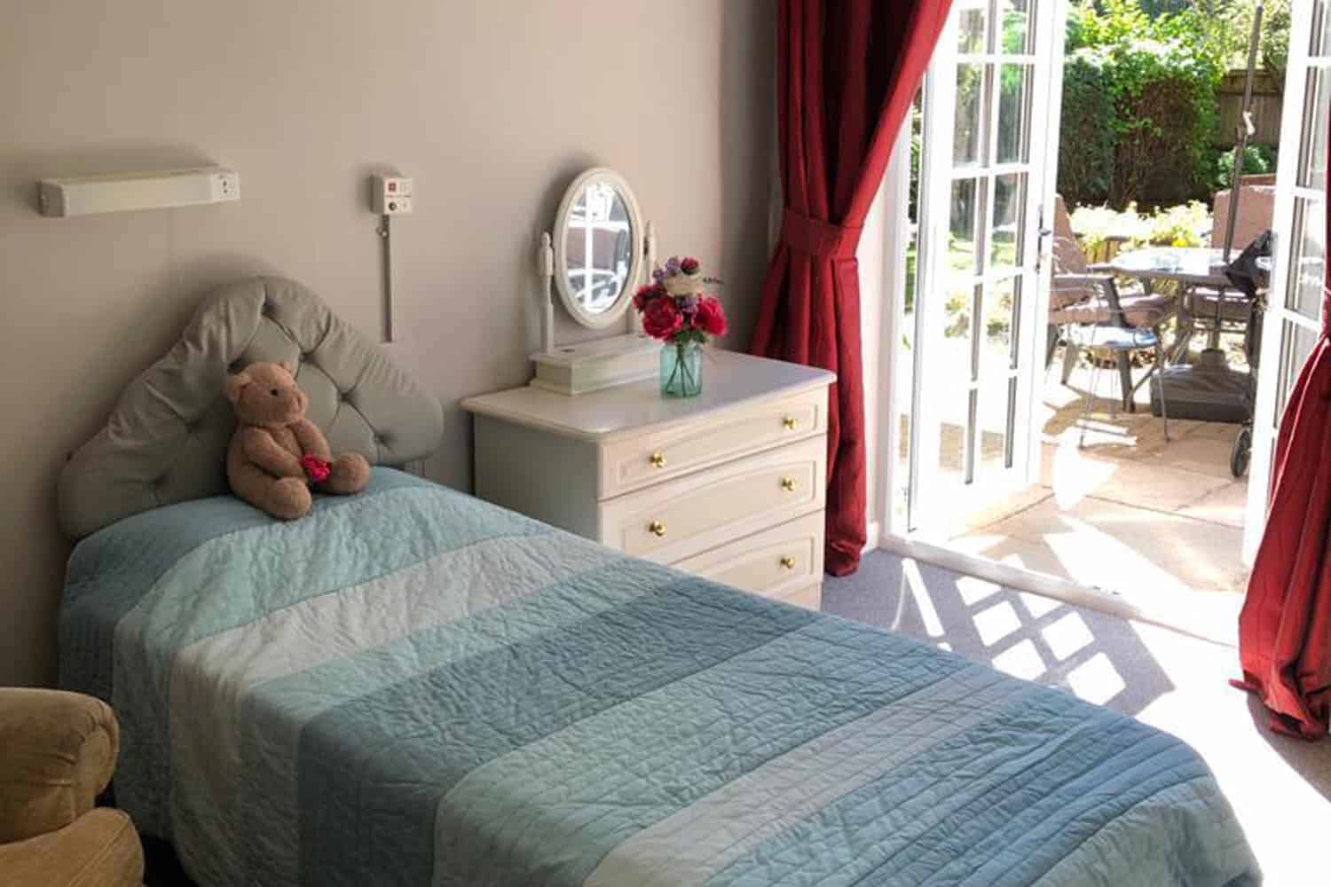 A cozy accommodation bedroom with a single bed adorned with a blue quilt, a teddy bear perched on top of a white pillow, a bedside table with a small bouquet of flowers, and a mirror