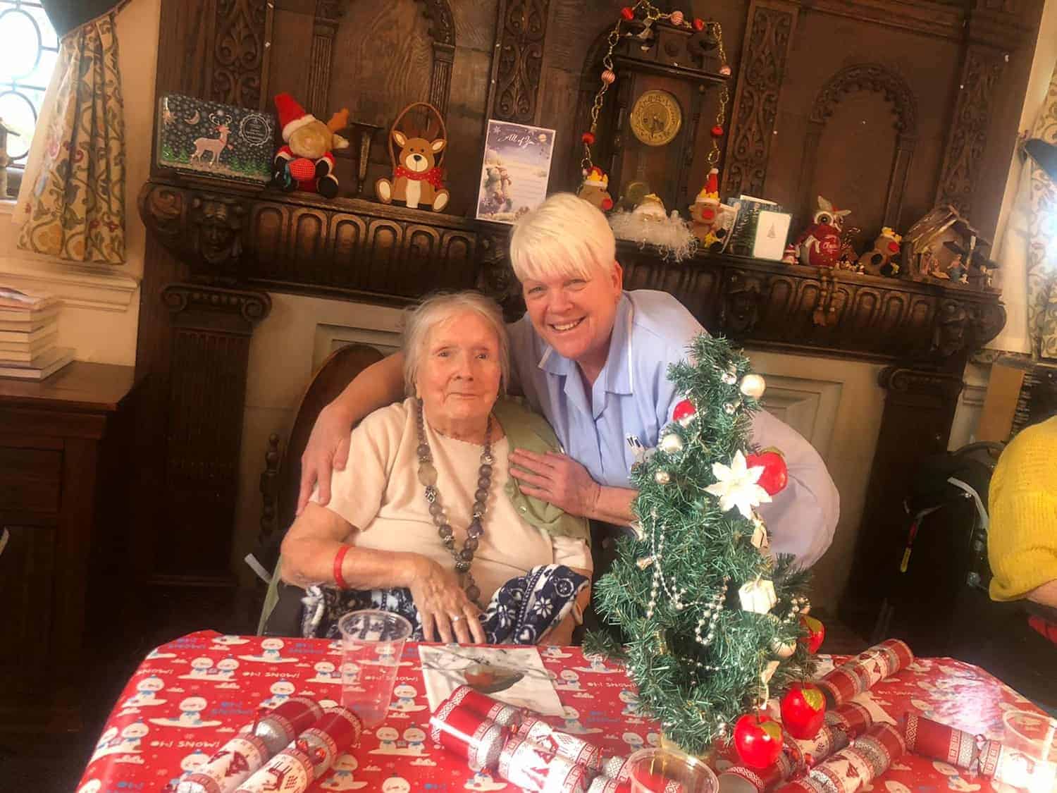 A joyful moment at a festive dinner table, with an elderly woman sitting beside a smiling caregiver, surrounded by christmas decorations, gifts, and a miniature christmas tree in a gallery setting, conveying a warm atmosphere