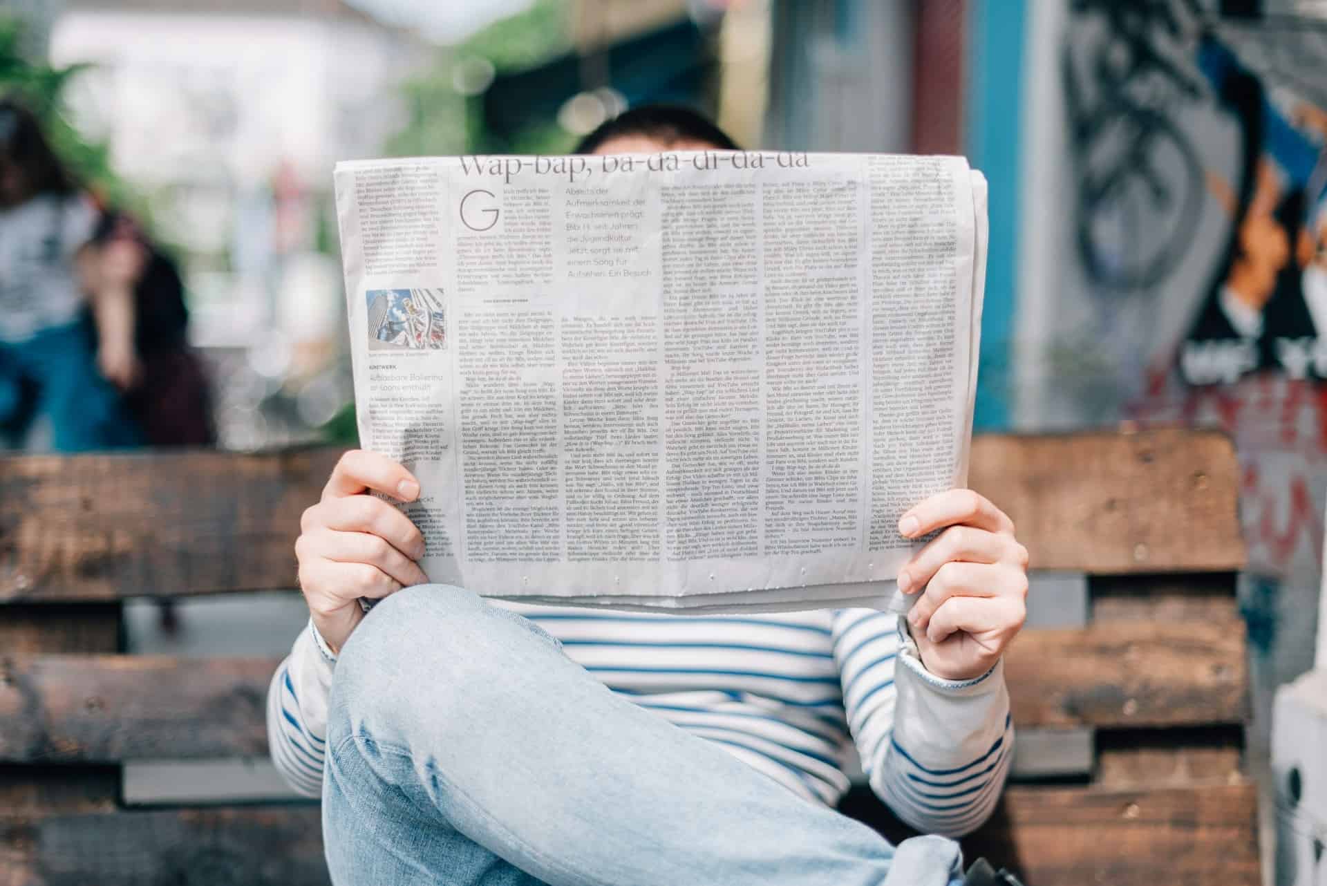 Person immersed in reading the latest news, concealing their face, in a casual urban setting.