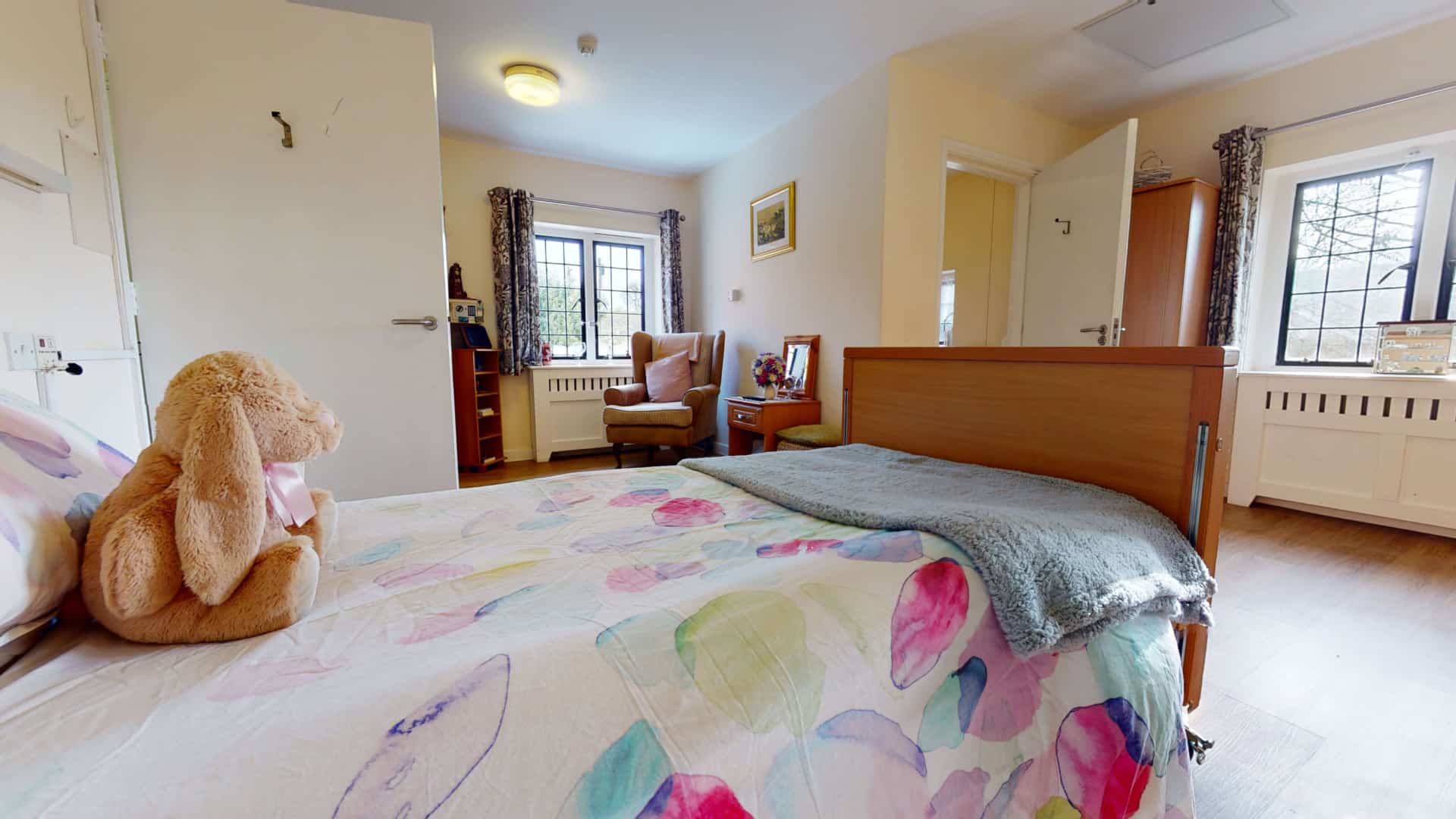 A cozy, well-lit bedroom in an assisted living facility featuring a large bed with a floral comforter, a giant plush bunny on the bed, an armchair by the window, and wooden furniture